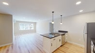 This thoughtfully designed single-floor residence offers a bright and contemporary living experience. The open-concept layout seamlessly connects the kitchen and living area, creating a spacious and functional environment. The kitchen serves as the centerpiece of the space, showcasing a large island with a dark granite countertop, an undermount sink, and pendant lighting above. White shaker-style cabinetry with matte black hardware lines the walls, complemented by matching dark granite counters. A full suite of stainless steel appliances — including a refrigerator, electric range, built-in microwave, and dishwasher — completes the well-equipped kitchen. Light hardwood flooring extends throughout the entire unit, lending warmth and continuity to each room. The living area benefits from natural light through large windows with outdoor views. Bedrooms are generously sized, with one featuring a walk-in closet and a ductless mini-split climate control unit. The bathroom features a white vanity with dark granite countertop, an undermount sink with a modern faucet, and a framed black mirror. Recessed lighting and neutral-toned walls throughout contribute to a clean, move-in-ready aesthetic.