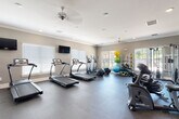 This fitness center is designed to accommodate a wide range of workout routines within a spacious, single-story layout. The interior features neutral gray walls, white crown molding, and recessed lighting complemented by ceiling fans, creating a clean and comfortable environment. Rubber flooring throughout provides durability and safety across all exercise zones.

The main cardio area houses multiple SportsArt treadmills and elliptical machines arranged along windowed walls fitted with white blinds, allowing ample natural light to fill the space. Flat-screen televisions are mounted at convenient viewing angles above the cardio equipment. A dedicated cycling area features several stationary spin bikes alongside additional SportsArt upright machines.

A separate free weights zone includes a full dumbbell rack, colorful kettlebell sets displayed on tiered racks, and a weight bench. Floor-to-ceiling mirrors line this area, enhancing the sense of space and allowing users to monitor their form. Nearby, a cable resistance machine offers additional strength training options.

A functional training station is stocked with medicine balls, stability balls of varying sizes, and resistance bands neatly organized on a dedicated rack. A water cooler station and posted fitness center guidelines are also present, adding practical convenience to the overall facility.