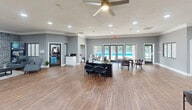 Located at 501 NC-54 W in Carrboro, NC, this single-floor commercial space offers a generously sized, well-appointed environment designed for both productivity and comfort. The open-plan main area features warm hardwood-style flooring, recessed lighting, and large glass entry doors that flood the space with natural light and provide views of the surrounding greenery. Multiple workstations are arranged throughout the open floor, complemented by a ceiling fan and textured ceilings that add a residential warmth to the professional setting.

The living room lounge area serves as a standout feature, anchored by a dramatic full-height brick fireplace wall with a mounted flat-screen television. Comfortable seating, a coffee table, and a lush green wall panel create an inviting, relaxed atmosphere ideal for meetings or breaks.

The fully equipped kitchen is thoughtfully designed with white shaker cabinetry, a subway tile backsplash, dark countertops, and stainless steel appliances including a refrigerator, range, and microwave. A breakfast bar with pendant lighting and bar stools provides additional seating. Windows throughout the kitchen offer pleasant outdoor views, enhancing the bright and airy feel of the entire space.