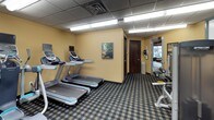 This fitness center is a fully outfitted exercise facility spanning a single level, designed to accommodate a wide variety of workout routines. The space features a drop ceiling with recessed fluorescent lighting that provides bright, even illumination throughout, while a patterned carpet in a classic plaid design covers the floor, adding a distinct character to the room.

The cardio section is equipped with multiple treadmills and elliptical machines, each fitted with individual monitor screens, allowing users to track their progress or enjoy entertainment during their workout. A rowing machine rounds out the aerobic equipment selection.

The strength training area is equally well-appointed, featuring a Precor cable machine system, a dedicated weight bench, and a full dumbbell rack offering a range of free weights. Additional Precor resistance machines provide targeted muscle training options for users of all fitness levels.

The walls are painted in warm yellow and earthy orange tones, accented by a teal feature wall, creating an energetic yet welcoming atmosphere. Decorative artwork is displayed throughout the space. A mirrored wall in the strength training section enhances the sense of openness. The facility includes a security camera and easy access through a dark wood-finished door, ensuring both safety and convenience for all users.