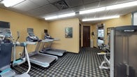 This fitness center is a fully outfitted exercise facility spanning a single level, designed to accommodate a wide variety of workout routines. The space features a drop ceiling with recessed fluorescent lighting that provides bright, even illumination throughout, while a patterned carpet in a classic plaid design covers the floor, adding a distinct character to the room.

The cardio section is equipped with multiple treadmills and elliptical machines, each fitted with individual monitor screens, allowing users to track their progress or enjoy entertainment during their workout. A rowing machine rounds out the aerobic equipment selection.

The strength training area is equally well-appointed, featuring a Precor cable machine system, a dedicated weight bench, and a full dumbbell rack offering a range of free weights. Additional Precor resistance machines provide targeted muscle training options for users of all fitness levels.

The walls are painted in warm yellow and earthy orange tones, accented by a teal feature wall, creating an energetic yet welcoming atmosphere. Decorative artwork is displayed throughout the space. A mirrored wall in the strength training section enhances the sense of openness. The facility includes a security camera and easy access through a dark wood-finished door, ensuring both safety and convenience for all users.