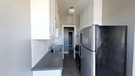 This single-floor space showcases a well-appointed galley-style kitchen that serves as a central highlight. The kitchen is designed for efficiency and style, featuring crisp white upper and lower cabinetry that extends to the ceiling, offering generous storage throughout. A classic white subway tile backsplash lines the walls, lending a timeless and clean aesthetic to the space. The countertops are finished in a cool gray tone, providing a sleek, modern contrast to the white cabinetry above. Stainless steel appliances, including a full-size refrigerator and a range, bring a contemporary, professional-grade feel to the kitchen. A single-basin sink with an elegant bridge-style faucet is neatly integrated into the countertop. The flooring features dark wood-tone planks that ground the space with warmth and visual depth. A classic schoolhouse pendant light fixture mounted to the ceiling provides focused, ambient lighting that complements the overall aesthetic. A doorway at the end of the kitchen leads further into the home, hinting at additional living spaces beyond. The overall ambiance is clean, functional, and stylishly understated, balancing classic design elements with modern conveniences.