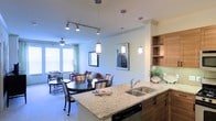 This single-floor residence showcases a thoughtfully designed open-concept layout that integrates the kitchen, dining, and living areas into one cohesive, flowing space. The kitchen is appointed with light granite countertops, a stainless steel sink, warm wood cabinetry with open upper shelving, and a green glass tile backsplash that adds a subtle pop of color. A peninsula counter extends into the dining area, offering additional workspace and a natural transition between cooking and entertaining zones. The dining area features a round dark-wood table surrounded by striped upholstered chairs, lit elegantly by a series of pendant lights that hang at varying heights, creating visual interest overhead. Track lighting further enhances the brightness of the space. The living area beyond is comfortable and well-lit, benefiting from large windows dressed with blinds and green curtains that allow ample natural light to filter in. A ceiling fan adds both function and style to this relaxed gathering space. Framed artwork on the walls and carefully chosen furnishings throughout contribute to the warm, residential ambiance. The seamless flow between all areas makes this space ideal for both everyday living and entertaining.