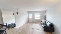 This single-floor unit offers a thoughtfully laid-out interior finished in a soft, neutral color palette throughout. The living area is a welcoming space, characterized by light gray walls, wood-look laminate flooring, and a distinctive white grid-panel accent wall that adds subtle architectural interest. Natural light enters through a front-facing window alongside a paneled entry door, while a wall-mounted air conditioning unit ensures year-round comfort. A large leaning mirror amplifies the sense of space, and hanging woven planters contribute a decorative touch.

The living area flows openly toward the kitchen, which is equipped with white cabinetry, speckled quartz-style countertops, a double stainless steel sink with a modern pull-down faucet, and a window above the counter for added natural light. A compact dining area accommodates a bistro-style table and two bar stools, making it ideal for casual dining.

The bedroom is a quiet, carpeted retreat with soft gray walls, ample natural light from a window with blinds, and direct access to the rest of the unit. The full bathroom features a tub-shower combination, a white vanity with matching countertop, and wood-look flooring, maintaining the consistent clean aesthetic found throughout the space.