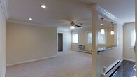 This single-floor space presents an open, airy layout designed for comfortable everyday living. The main living area is finished with plush wall-to-wall carpeting and neutral-toned walls, creating a warm and inviting atmosphere. Crown molding runs along the ceiling perimeter, adding a refined architectural detail throughout the space. A ceiling fan with an integrated light fixture is centrally positioned, providing both air circulation and ambient lighting, complemented by recessed ceiling lights that enhance overall brightness.

A decorative column with a classical capital defines the boundary between the living area and an adjacent kitchen space, which features dark countertops and a pendant light fixture overhead, hinting at a functional and stylish cooking area. Natural light flows in through multiple windows fitted with white blinds, keeping the space bright while offering privacy. A wall-mounted HVAC unit is positioned beneath the windows, ensuring climate control. A paneled interior door leads to an additional room, likely a bedroom or storage area, further adding to the functional layout of this well-appointed space.