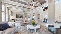 This beautifully designed single-floor open-concept space offers a generous footprint that seamlessly integrates multiple functional zones into one cohesive, light-filled environment. The architectural design is immediately impressive, with dramatically soaring ceilings accented by exposed wood-paneled coffered beams and a dark wood inset ceiling detail that adds warmth and visual depth.

The main living area is anchored by a large, plush area rug and sophisticated neutral-toned seating arranged around a whitewashed octagonal coffee table. A striking shiplap accent wall serves as the focal point, housing a sleek linear fireplace topped by large-scale coastal artwork, with a marble-toned hearth ledge adding an elegant finish. Multiple modern globe and cluster chandeliers illuminate the space with a contemporary yet inviting glow.

Floor-to-ceiling windows along one wall flood the interior with natural light and offer expansive outdoor views, enhancing the open, airy ambiance. Light-toned wide-plank flooring runs throughout, tying the zones together cohesively.

A large kitchen island with bar-height seating provides a gathering hub, complemented by a textured stone backsplash accent wall in the adjoining wet bar or catering area. A separate dining zone with ample seating and additional pendant lighting rounds out this well-appointed space, making it ideal for entertaining and social gatherings.