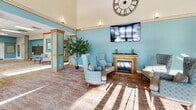 Located at 434 W 4th St in Red Wing, Minnesota, this single-floor community facility offers a generous and thoughtfully designed layout across its expansive floor plan. The centerpiece is a dramatic main lounge area distinguished by soaring two-story ceilings, teal accent walls, warm wood-trimmed columns, and abundant natural light streaming through large windows. An elegant gold-framed fireplace anchors the seating area, complemented by a mounted flat-screen television and a large decorative Roman numeral clock overhead. Comfortable upholstered armchairs and sofas invite residents and visitors to gather and relax. A wide central corridor connects the various areas of the facility, lined with a built-in mailbox wall, seating nooks, decorative artwork, and indoor greenery that lend warmth throughout. The activity and multipurpose room features a drop ceiling with recessed panel lighting, movable tables and chairs, a wall-mounted display screen, and a projector — ideal for events and programming. An adjacent full kitchen offers warm oak cabinetry, a large center island, full appliances, and tile flooring. A dedicated library and reading area houses well-stocked open bookshelves, cozy seating, and a writing desk with exterior-facing windows. A compact resource office provides a workstation and informational displays for residents.
