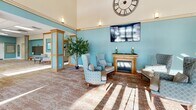 Located at 434 W 4th St in Red Wing, Minnesota, this single-floor community facility offers a generous and thoughtfully designed layout across its expansive floor plan. The centerpiece is a dramatic main lounge area distinguished by soaring two-story ceilings, teal accent walls, warm wood-trimmed columns, and abundant natural light streaming through large windows. An elegant gold-framed fireplace anchors the seating area, complemented by a mounted flat-screen television and a large decorative Roman numeral clock overhead. Comfortable upholstered armchairs and sofas invite residents and visitors to gather and relax. A wide central corridor connects the various areas of the facility, lined with a built-in mailbox wall, seating nooks, decorative artwork, and indoor greenery that lend warmth throughout. The activity and multipurpose room features a drop ceiling with recessed panel lighting, movable tables and chairs, a wall-mounted display screen, and a projector — ideal for events and programming. An adjacent full kitchen offers warm oak cabinetry, a large center island, full appliances, and tile flooring. A dedicated library and reading area houses well-stocked open bookshelves, cozy seating, and a writing desk with exterior-facing windows. A compact resource office provides a workstation and informational displays for residents.