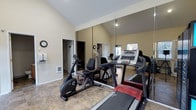 This single-floor fitness space is thoughtfully designed to serve as a fully functional workout room. The room immediately impresses with its vaulted ceilings, which lend an open, airy feel and make the space feel larger than its footprint suggests. A full-wall mirror runs along one side of the room, visually expanding the space further while allowing users to monitor their form during exercise sessions.

The fitness area is equipped with a lineup of cardio machines, including a treadmill and a recumbent stationary bike, making it well-suited for a variety of cardiovascular workouts. A weight bench adds versatility, supporting strength training in addition to cardio activities. The flooring features a neutral stone-look tile that is both durable and easy to maintain.

Warm, neutral-toned walls create a calm and focused atmosphere, while a wall clock ensures users can keep track of their workout time. A window allows natural light to filter in, contributing to a bright and energizing environment. A doorway connects the fitness room to adjacent areas of the space, ensuring convenient accessibility. The overall design balances functionality with comfort, making this a practical and inviting space for daily fitness routines.