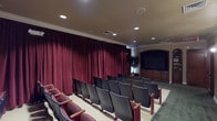 This single-floor screening room is designed to deliver a dedicated, theater-style viewing experience in a refined setting. The room is arranged with multiple rows of individual theater seats featuring burgundy upholstered cushions and dark armrests, organized in a tiered layout to ensure clear sightlines to the front display area. Floor-to-ceiling burgundy drapes line the walls, providing acoustic dampening and a classic cinematic aesthetic that reinforces the room's purpose.

At the front of the room, a large flat-panel display screen is set within a built-in wood-paneled cabinetry unit finished in warm tones. Flanking cabinetry includes additional storage and display shelving, adding a polished, furnished quality to the viewing wall. A decorative potted tree accents the front corner, introducing a natural element to the otherwise structured interior.

Recessed ceiling lighting provides even, adjustable illumination throughout the seating area, while crown molding along the ceiling perimeter contributes an architectural refinement to the space. A rear entry area features rich wood double doors, wall sconces, and green carpeting, serving as a transitional foyer before entering the main screening room. The overall design balances functional entertainment infrastructure with a sophisticated, well-appointed interior finish.