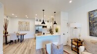 This thoughtfully designed single-floor residence offers a cohesive blend of modern finishes and functional living spaces. The heart of the home is a striking open-concept kitchen, showcasing crisp white shaker cabinetry with glass-front upper cabinets, a quartz waterfall island, stainless steel appliances including a French door refrigerator and range with a sleek hood, and matte black fixtures throughout. A pair of industrial-style pendant lights adds a warm, contemporary touch above the island. Light-toned hardwood flooring flows seamlessly from the kitchen into the dining and living areas, creating a bright and cohesive aesthetic. The dining area accommodates a solid wood table and is accented by nature-inspired artwork. The living room features a ceiling fan, large-format abstract wall art, and sliding glass doors that open onto a private balcony, filling the space with natural light and outdoor views. The bedroom is a calm retreat, carpeted for comfort and equipped with a walk-in closet and a tray ceiling detail. The full bathroom is finished in large-format tile, paired with white cabinetry, quartz countertops, matte black hardware, and a full tub-shower combination — maintaining the clean, modern palette found throughout the entire residence.
