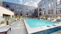 Located at 50 S Grant Ave in Columbus, Ohio, this outdoor amenity space is set within a modern multi-story building complex featuring a striking gray panel and mixed-material facade with large windows and private balconies. The centerpiece of the space is a generously sized, resort-style swimming pool with vivid turquoise water and decorative water jets, framed by wide, large-format tile pool decking. Surrounding the pool, rows of contemporary wood-and-white cushion chaise lounges provide ample space for sunbathing, all set against the architectural backdrop of the enclosing building wings. Glass panel railings define the tiered levels of the outdoor space, lending an open, unobstructed sightline throughout. A separate lounge terrace features plush sectional seating, curated landscaping with ornamental plantings, and a fire table, creating an intimate gathering area distinct from the pool deck. Mature trees and seasonal plantings are integrated throughout, softening the urban setting. The layout is thoughtfully designed to maximize open-air enjoyment, with clear sightlines across all zones and access points connecting the pool deck, spa, and lounge areas seamlessly.