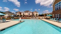 Located at 1100 Deer Harbor Dr in Garner, NC, this outdoor amenity area centers around a generously sized, resort-style swimming pool with crystal-clear blue water. The pool deck is thoughtfully designed with a wide concrete surround, accommodating numerous lounge chairs and umbrella-shaded seating arrangements for sunbathing and relaxation. A covered cabana structure, supported by clean white columns, provides a shaded retreat directly adjacent to the pool. This covered area is furnished with comfortable striped cushioned seating and additional lounge chairs, making it an ideal gathering spot. The pool area is enclosed by a black wrought-iron fence, with a retaining wall lined with manicured landscaping and seasonal plantings adding color and structure to the perimeter. A dedicated pool house building, featuring a combination of brick and lap siding with a navy blue entry door, provides access to amenities and displays posted pool rules for resident safety. The backdrop includes views of multi-story residential buildings framed by mature trees, reinforcing the community setting. Open to abundant natural light under expansive skies, this outdoor space offers a welcoming, well-appointed environment suited for both leisure and recreation.