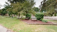 This inviting outdoor space is a well-maintained courtyard that blends natural beauty with functional design. Mature deciduous trees provide generous canopy coverage, casting dappled shade across the grounds and creating a cool, tranquil atmosphere. The lawn is neatly kept, with a defined mulched seating area serving as the focal point of the space. Two classic wrought-iron park benches sit side by side on the mulched platform, offering a quiet spot for rest or conversation. Nearby, a traditional round picnic table with attached seating provides an additional area ideal for casual outdoor gatherings or al fresco dining. Lush, dense shrubs border the seating area, adding layers of greenery and a sense of enclosure that enhances the sense of privacy. In the background, a low-profile building facade is partially visible through the foliage, suggesting the courtyard serves as an accessible outdoor extension of an adjacent structure. The overall ambiance is calm and park-like, making this space well-suited for leisurely breaks, social interaction, or quiet reflection in a green, natural setting.