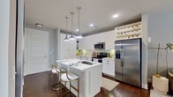This single-floor residence showcases a thoughtfully designed open-concept layout that seamlessly connects the kitchen, dining, and living areas. The kitchen is a standout feature, outfitted with crisp white cabinetry extending to the ceiling, a subway tile backsplash, stainless steel appliances including a French door refrigerator and electric range, and a large center island with a waterfall-edge countertop. Three industrial-style pendant lights hang above the island, which offers seating for casual dining. A built-in wine rack adds a refined touch to the upper cabinetry. Dark hardwood flooring flows throughout, providing a warm contrast to the light finishes.

The living area receives generous natural light through large windows dressed with horizontal blinds, creating a bright and airy atmosphere. The bedroom is comfortably sized with a wide window, dark dresser, and room for expressive artwork.

The bathroom is elegantly appointed with a full-length double vanity featuring white cabinetry, a soaking tub, and a large walk-in tiled shower. The combination of dark wood flooring, grey tile walls, and a wide mirror contributes to a clean, spa-like environment throughout.