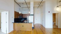 This single-floor loft offers a compelling blend of industrial character and modern finishes. The open-concept layout is immediately striking, with soaring ceilings that amplify the sense of space and light throughout. Original wide-plank hardwood floors run underfoot, lending warmth and texture to the interior.

A defining architectural feature is the prominent white-tiled structural column, capped with a decorative green-glazed ceramic detail — a nod to the building's historic character. This column anchors the transition between the living area and the kitchen, serving as both a functional and visual centerpiece.

The kitchen is well-appointed with warm wood cabinetry, bold dark granite countertops, and a classic dark tile backsplash. Stainless steel appliances, including a range, microwave, and refrigerator, complete the modern culinary setup. A peninsula extends from the column, providing additional prep and seating space.

Industrial-style exposed ceiling tracks with adjustable spotlights provide flexible lighting throughout, complementing the loft's raw aesthetic. Pendant lights hang above the kitchen area, adding a refined touch. Large windows allow natural light to filter in, brightening the open living space. The overall ambiance is urban and contemporary, with historic architectural details that give the space a distinctive, one-of-a-kind character.