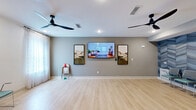 Located at 720 Verdae Blvd in Greenville, SC, this single-floor fitness and wellness studio offers a well-designed space ideal for group classes and individual training sessions. The main open floor area is finished with light hardwood-style flooring throughout, creating a clean, contemporary aesthetic that is both functional and inviting.

The primary exercise area is generously proportioned, featuring a large wall-mounted flat-screen TV flanked by framed artwork, making it well-suited for instructor-led or virtual fitness classes. Two sleek ceiling fans provide airflow across the open floor, enhancing comfort during active use. Natural light filters in through draped windows along one wall, contributing to a bright and energizing atmosphere.

A striking accent wall adorned with a bold blue chevron-patterned wallcovering lines one side of the room, in front of which a row of teal upholstered chairs provides seating for participants. Adjacent to the main floor, a dedicated equipment area houses stability balls, dumbbells, medicine balls, and other fitness accessories neatly organized on racks.

The entry area includes a water station, cabinetry, and access to additional rooms, ensuring a complete and practical layout. Recessed lighting throughout maintains a polished, professional ambiance, making this studio a versatile and well-appointed facility for a variety of fitness and wellness programming.