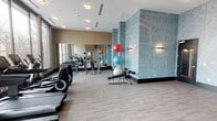 This contemporary fitness center is thoughtfully designed to support a full range of workout routines in a clean, modern environment. The space is organized into distinct functional zones, allowing for both cardio training and strength work without congestion. Large floor-to-ceiling windows line one side of the facility, flooding the cardio area with abundant natural light and offering views of the surrounding streetscape — an energizing backdrop for treadmills, ellipticals, upright bikes, and a stair climber, all from Life Fitness. The light wood-look flooring throughout contributes to a bright, welcoming atmosphere. A defining design feature is the prominent accent wall clad in a soft blue-toned city map graphic, adding a bold visual identity to the space and complementing the neutral beige and warm wood tones used elsewhere. The strength and functional training area is well-appointed with a multi-use cable machine, adjustable weight bench, resistance bands, kettlebells, and a tiered rack holding stability balls in multiple sizes. Full-length mirrors expand the visual depth of this zone and allow users to monitor their form. A water fountain station is conveniently integrated into the layout. Recessed and flush ceiling lighting ensures consistent illumination throughout, while wall-mounted sconces add a refined touch to the accent wall area.
