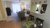 This single-story residence showcases an open-concept layout that seamlessly connects the dining and living areas, creating a cohesive and functional living environment. The dining area features hardwood-style flooring and is furnished with a glass-top table and black chairs, providing a clean, modern contrast to the warmer tones throughout the space.

A breakfast bar with bar stools serves as a natural divider between the dining area and the adjacent kitchen, which is partially visible through a pass-through window. This design element enhances the open feel while maintaining a defined sense of each zone.

The living area transitions to plush carpeting and is anchored by a comfortable seating arrangement. A ceiling fan with a light fixture overhead provides both air circulation and ambient lighting. Large vertical blinds frame a sliding glass door that floods the room with natural light and offers access to an outdoor area.

A mirror on the wall reflects additional light and provides a glimpse into a nearby bedroom, adding depth to the space. The overall ambiance is warm and inviting, with neutral tones and layered lighting creating a comfortable, livable atmosphere throughout.