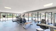 Located at 1701 37th Street in Phenix City, Alabama, this fitness center offers a thoughtfully designed, single-floor workout facility. The space is organized to accommodate a wide range of exercise needs, with dedicated zones for both cardio and strength training. Along the front of the room, expansive floor-to-ceiling glass windows and sliding doors flood the interior with natural light and provide unobstructed views of an outdoor pool area, lush greenery, and the surrounding property — creating an energizing atmosphere for workouts. The cardio section is well-appointed with multiple treadmills, elliptical trainers, and stationary bikes positioned to take full advantage of the outdoor views. Toward the rear, the strength and functional training area features a cable machine, weight bench, free weights, and a step platform. A full-length mirror with a wooden ballet barre runs along one interior wall, offering additional space for stretching, balance training, or barre-style exercises. Motivational and instructional posters are mounted on the walls. The flooring throughout is durable, cushioned gym matting in a neutral gray tone, providing traction and support. Recessed panel lighting in the ceiling ensures consistent, even illumination across the entire space. The overall design is clean, modern, and functional.