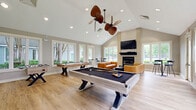 Located at 2018 Cunningham Dr in Hampton, Virginia, this well-appointed community recreation room offers a welcoming and versatile gathering space designed for both relaxation and entertainment. The room is anchored by dramatic vaulted ceilings that rise to a peak, creating an open, airy atmosphere enhanced by recessed lighting and dual tropical-style ceiling fans that add a casual, resort-like charm.

Light hardwood flooring runs throughout the single-level space, complementing the neutral warm-gray wall tones and crisp white trim. A full-size pool table with a sleek dark felt surface serves as the centerpiece of the room, accompanied by a shuffleboard table for additional recreation. Adjacent to the game area, a comfortable lounge zone features tufted leather sofas arranged around a patterned area rug and a dark coffee table, all positioned near a modern fireplace with a wall-mounted television above it.

An abundance of windows on multiple walls floods the interior with natural light and offers views of the surrounding greenery. A pass-through window connects to a small kitchenette area, adding functional convenience. Multiple entry points, including glass-paneled doors, provide easy access and further enhance the sense of openness. This space is thoughtfully designed to serve as a dynamic hub for socializing and leisure.