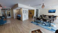 This single-floor space offers an open, well-organized layout that combines multiple functional areas within a bright and welcoming environment. The flooring throughout is a warm hardwood-style material that ties the space together cohesively, providing both durability and visual appeal.

The main gathering area is anchored by a large dining table surrounded by dark metal-framed chairs, set atop a geometric blue area rug that adds a pop of color and texture. A sculptural pendant light with a layered wood design hangs above, serving as a striking focal point. Adjacent to the dining area, a cozy display and activity nook features decorative shelving with subtle accent lighting, adding a creative and personalized touch.

A central dividing wall, accented with a bold mustard-yellow paint, helps define zones within the open floor plan while displaying framed artwork and a nautical anchor motif — lending the space a coastal character. Recessed lighting is installed throughout the ceiling, ensuring consistent and even illumination across all areas.

Large glass-paneled doors or windows near the rear allow for natural light to filter in, enhancing the airy feel of the space. The overall ambiance is casual yet thoughtfully styled, making this an inviting and functional environment suited for gathering, socializing, or everyday use.