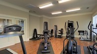 This fitness center offers a thoughtfully organized workout space designed to accommodate a range of exercise routines. The room features warm hardwood flooring throughout, neutral gray walls with crisp white crown molding and baseboards, and bright overhead fluorescent lighting that ensures a well-lit environment for training at any time of day. The space is outfitted with a selection of commercial-grade strength machines, including cable and resistance units positioned to allow comfortable use and adequate spacing between equipment. A dedicated free weight area includes a full dumbbell rack, providing options for a variety of strength training exercises. Cardio equipment, including a stationary bike and an elliptical machine, rounds out the fitness offerings. Protective floor mats are placed beneath the equipment, adding safety and durability underfoot. A doorway visible from the interior suggests a connected anteroom or entryway, contributing to the overall flow and accessibility of the facility. The clean, functional layout and quality equipment selection make this a practical and comprehensive fitness space suitable for a wide range of users and fitness levels.
