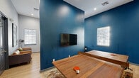 Located at 7801 Scenic Hwy in Baton Rouge, Louisiana, this single-floor game room and lounge delivers an energetic, social atmosphere designed for recreation and entertainment. The space is defined by a striking bold blue accent wall that anchors the room and creates a dynamic contrast against the surrounding light gray walls and light wood-toned flooring.

At the center of the space sits a full-sized ping-pong table crafted from rich hardwood, serving as the focal point of the main game area. A wall-mounted flat-screen TV is positioned on the blue accent wall, complemented by a wood-toned media console flanked by potted snake plants, adding a touch of greenery. Natural light filters in through multiple windows fitted with white horizontal blinds.

A dedicated poker area features a round gaming table surrounded by upholstered chairs, set against the deep blue walls and accented by large playing card-themed artwork. Adjacent seating includes a pair of cognac leather lounge chairs arranged around a small side table, ideal for casual conversation.

The lounge area opens through glass-paned doors into a neighboring space furnished with high-top bistro tables and stools, its walls adorned with colorful, whimsical robot-themed artwork panels. Recessed ceiling lighting throughout ensures the space is well-lit, enhancing its lively and inviting ambiance.