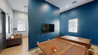 Located at 7801 Scenic Hwy in Baton Rouge, Louisiana, this single-floor game room and lounge delivers an energetic, social atmosphere designed for recreation and entertainment. The space is defined by a striking bold blue accent wall that anchors the room and creates a dynamic contrast against the surrounding light gray walls and light wood-toned flooring.

At the center of the space sits a full-sized ping-pong table crafted from rich hardwood, serving as the focal point of the main game area. A wall-mounted flat-screen TV is positioned on the blue accent wall, complemented by a wood-toned media console flanked by potted snake plants, adding a touch of greenery. Natural light filters in through multiple windows fitted with white horizontal blinds.

A dedicated poker area features a round gaming table surrounded by upholstered chairs, set against the deep blue walls and accented by large playing card-themed artwork. Adjacent seating includes a pair of cognac leather lounge chairs arranged around a small side table, ideal for casual conversation.

The lounge area opens through glass-paned doors into a neighboring space furnished with high-top bistro tables and stools, its walls adorned with colorful, whimsical robot-themed artwork panels. Recessed ceiling lighting throughout ensures the space is well-lit, enhancing its lively and inviting ambiance.