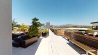 This expansive rooftop terrace offers a well-designed outdoor retreat with sweeping panoramic views of the surrounding mountains and cityscape under open skies. The deck is finished with large-format light gray tiles that lend a clean, contemporary aesthetic throughout the space. Thoughtfully zoned seating areas are defined by horizontal wood-slat privacy partitions, creating intimate pockets for relaxation and socializing while maintaining the openness of the terrace. Substantial sculptural fire bowls in a rich, dark metallic finish serve as dramatic focal points, complemented by additional decorative planters in both matte black and weathered corten-style materials, filled with bamboo and ornamental grasses that add a natural, verdant contrast to the modern hardscape. A wall-mounted outdoor television is positioned against the clean, panel-clad exterior wall, providing an entertainment option alongside the striking natural backdrop. Wood-and-steel benches are distributed throughout for casual seating, and a dedicated entrance with a glass door provides rooftop access. The overall ambiance is sophisticated and resort-like, combining modern architectural elements with abundant greenery and unobstructed views to create an inviting open-air amenity space.