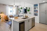 This thoughtfully designed single-floor residence at 1901 E Dyer Rd in Santa Ana, California offers a cohesive open-concept layout that seamlessly connects the kitchen, dining, and living areas. The kitchen is a standout feature, showcasing custom gray shaker cabinetry with brushed gold hardware, pristine white quartz countertops, a large center island with an undermount sink, and a full suite of stainless steel appliances including a gas range and French door refrigerator. A vibrant blue-tiled built-in shelving unit adds a distinctive design accent. Pendant lighting illuminates the island, while recessed lighting throughout ensures the space remains bright and welcoming. Light hardwood flooring flows continuously across the main living areas, enhancing the sense of openness. The living area benefits from generous natural light through large windows, offering views of the surrounding urban landscape, and is anchored by textured accent walls and lush greenery. The dining nook features a round marble-top table set beside additional windows. The bedroom is serene and well-appointed, with a patterned ceiling treatment, wall-mounted sconces, a natural rattan bed frame, and teal curtains that frame an oversized window. The bathroom completes the space with matching gray cabinetry, gold accents, a quartz vanity, and a tiled shower enclosure.