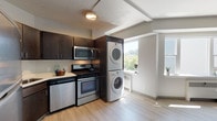 Located at 707 West 5th Avenue in Spokane, Washington, Unit 1109 is a thoughtfully designed single-floor residence that balances functionality with clean, modern aesthetics. Light-toned luxury vinyl plank flooring runs throughout the entire unit, creating a cohesive and easy-to-maintain living environment.

The open living area is bright and airy, benefiting from large windows that frame views of the surrounding tree canopy and fill the space with natural light. Exposed ceiling beams add a subtle architectural character to the room.

The kitchen is efficiently laid out in an L-shaped configuration, featuring dark espresso cabinetry, stainless steel appliances including a refrigerator, range, and over-the-range microwave, as well as a stainless steel sink. A standout convenience is the in-unit stacked washer and dryer, positioned adjacent to the kitchen area and illuminated by its own window.

The bedroom is a generously sized, quiet room with access to a dedicated closet, providing ample storage. The bathroom is neatly appointed with light-toned flooring, a pedestal-style vanity, a toilet, and a glass-door walk-in shower stall. The overall design of the unit prioritizes practicality and comfort, making it a well-rounded urban residence in the heart of Spokane.