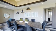 This contemporary conference room offers a well-appointed setting for professional meetings and collaborative gatherings. At the heart of the space sits a bold, dark wood-grain conference table surrounded by sleek black ergonomic chairs, providing both comfort and a polished aesthetic. Three eye-catching matte black pendant lights with gold interiors hang above the table, casting a warm, focused glow that complements the room's neutral color palette of soft whites and warm beige tones. Recessed ceiling lighting adds to the bright, professional atmosphere, while a distinctive dark accent beam frames the upper perimeter of the space, adding architectural depth. The walls are adorned with a pair of modern abstract artworks featuring soft teal and white tones, contributing a refined yet approachable ambiance. Small potted greenery arrangements placed along the center of the table introduce a natural element, softening the room's otherwise structured design. The overall aesthetic strikes a balance between modern sophistication and functional practicality, making this conference room a versatile space well-suited for business meetings, presentations, and team discussions.