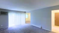 This single-floor space features a clean, functional layout with soft carpeted flooring running throughout the main living area and into the adjoining room. The living area is illuminated by natural light streaming through a large sliding glass door dressed with white vertical blinds, which also provides potential access to an outdoor area. Along the base of the wall, a white baseboard heater offers an efficient heating solution. A wide doorway connects the main living space to an adjacent room, which appears to be finished in warm neutral tones and features its own window and baseboard heating unit, ensuring consistent comfort throughout. The walls are painted in a cool, soft gray-blue palette, lending the space a calm and understated ambiance. The overall layout is open and straightforward, with clean white trim and minimal architectural detail creating a practical, move-in-ready environment. The abundance of natural light and neutral finishes provide a versatile backdrop suitable for a variety of furnishing styles.