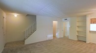 This two-floor residential space offers a functional and straightforward layout designed for comfortable everyday living. The main level features wall-to-wall carpeting that extends throughout the open living area, creating a warm and cohesive feel underfoot. A staircase with a white metal railing is prominently positioned along one wall, providing access to the upper floor while adding a clean, architectural element to the space. The textured popcorn ceiling adds a classic finish overhead, and neutral wall tones in soft white and warm gray contribute to a bright, understated ambiance. Natural light filters in through windows dressed with horizontal blinds, illuminating the interior and enhancing the sense of openness. A hallway connects the living area to additional rooms, with a glimpse of a carpeted secondary space visible in the background, suggesting a versatile floor plan. HVAC vents are integrated into both the ceiling and lower wall, ensuring climate control throughout the space. The overall design is clean and neutral, providing a blank canvas well-suited for a variety of furnishing styles and personal touches across both floors.