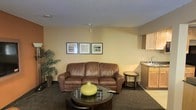 This single-floor space combines a living area and kitchenette into a cohesive, functional layout. The living area is anchored by a full-sized brown leather sofa positioned against warm, cream-toned walls adorned with a trio of framed artwork pieces, adding a touch of character to the space. A round, dark-finished coffee table sits in front of the sofa, styled with a decorative green vase, while a small side table nearby provides additional surface space. The carpeted flooring runs throughout the main area, contributing to a cozy atmosphere, and a potted tropical plant in the corner adds a natural, lively element. A textured ceiling completes the overhead detail. Tucked to one side, the kitchenette features light wood cabinetry and a compact sink, offering basic amenities within easy reach. The overall design is straightforward and practical, with neutral tones and understated décor creating a welcoming, no-frills environment suited for everyday living or short-term stays.