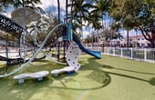 Located at 1140 SE 24th Rd in Homestead, Florida, this outdoor playground offers a well-appointed recreational space designed for active play in a sunny South Florida setting. The area is fully enclosed by a white metal fence, providing a secure environment for children and visitors alike.

The play area features a modern, multi-element playground structure that includes a climbing wall with hand and foot holds, a rope climbing dome, multiple slides in blue and teal, and spring-mounted balance steppers. The equipment is thoughtfully arranged to encourage both individual and group play. A separate seating area with green benches is positioned along the perimeter, offering comfortable spots for supervising adults.

The ground throughout the play area is covered with synthetic turf, providing a safe and cushioned surface underfoot. Towering palm trees frame the space, offering natural shade and lending a distinctly tropical character to the environment. Additional shade structures with decorative canopy tops are also present near the play equipment.

The playground benefits from abundant natural light and open skies, creating a bright and inviting atmosphere. Surrounding greenery, including hedges and mature trees along the fence line, enhances the sense of enclosure and privacy. The overall setting reflects a well-maintained community recreational space suited for children of varying ages.