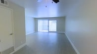 This single-floor space offers a clean, open layout designed to maximize both comfort and functionality. The main living area is immediately welcoming, characterized by soaring ceilings that enhance the sense of volume and openness throughout the room. A ceiling fan is centrally mounted, providing both practical airflow and a subtle design accent overhead.

The flooring throughout the living area consists of smooth, hardwood-style planks in a warm neutral tone, lending a polished and cohesive look to the space. The walls are finished in a soft, muted green-gray palette that contributes to a calm and understated ambiance.

A prominent sliding glass door spans a generous portion of one wall, dressed with vertical blinds that allow for adjustable light control. This feature serves as the primary source of natural light, brightening the interior and offering a visual connection to the outdoor area beyond. The door also provides convenient access to what appears to be a patio or exterior space.

White baseboards frame the perimeter of the room, offering a crisp contrast to both the flooring and the wall color. Overall, the space presents a bright, well-maintained, and versatile interior that is move-in ready and adaptable to a variety of furnishing styles and uses.