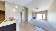 This single-floor space showcases an open-concept layout that integrates the kitchen and living areas into one cohesive environment. The vaulted ceilings rise dramatically, creating a strong sense of volume and airiness throughout the main living area. A ceiling fan is centrally mounted, providing both function and a finishing architectural detail.

The kitchen features a granite-topped breakfast bar with pendant lighting hanging above, offering a natural division between the cooking and living spaces without sacrificing the open feel. Dark cabinetry contrasts with the light countertop surfaces, adding visual depth.

The living area transitions from hardwood-style flooring to plush carpeting, clearly defining the lounge zone. A floor-to-ceiling tile-surround fireplace serves as a prominent focal point on the far wall, adding both warmth and a refined design element.

Large sliding glass doors flood the space with natural light and provide direct views to the exterior. The neutral warm-gray wall tones throughout the space complement the mixed flooring materials, contributing to a cohesive and modern aesthetic. An additional doorway visible from the main area suggests further rooms beyond.