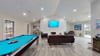 This single-floor space is designed with a distinct coastal aesthetic, blending modern finishes with relaxed, beach-inspired décor throughout. The centerpiece of the main living area is a dramatic vaulted shiplap accent wall, painted crisp white and anchored by a built-in fireplace at its base and a large wall-mounted flat-screen television above. Vaulted ceilings with recessed lighting add a sense of openness and volume to the space, while light gray wood-look flooring runs consistently throughout, tying each area together seamlessly.

The main gathering area is generously sized, accommodating a full seating arrangement with dark upholstered sofas and accent chairs arranged around a natural fiber area rug and a rustic coffee table. A full-size pool table occupies one side of the room, adding a recreational focal point. Sliding glass doors on the far end of the space allow abundant natural light to pour in and provide direct access to an outdoor pool area.

Adjacent to the main living area, a compact but fully equipped kitchen features white shaker cabinetry, subway tile backsplash, stainless steel appliances, and a double sink. Coastal artwork and decorative accents, including a decorative surfboard, reinforce the beach-inspired theme consistently across every area of this well-appointed space.