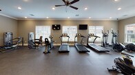 Located at 100 Legends Dr in Canton, GA, this fitness center offers a comprehensive, well-equipped workout facility designed to support a wide range of exercise routines. The space is thoughtfully divided into distinct workout zones, each catering to different fitness needs. The cardio area features multiple treadmills, elliptical trainers, and stationary bikes, including a Matrix cycling bike, all positioned in front of large windows with white plantation shutters that allow natural light to fill the room. Wall-mounted TVs and ceiling fans add to the comfort of the cardio zone. The strength and free weights area is anchored by a full dumbbell rack and a colorful kettlebell collection, both reflected in floor-to-ceiling mirrored walls that enhance the sense of space. Adjustable weight benches and a TRX cable machine round out the strength training options. A dedicated functional fitness section includes stability balls, resistance bands, and exercise mats, supported by additional cable and weight machines. The flooring transitions between rubber sport tiles and carpet, suitable for both high-impact and stationary exercises. Crown molding and neutral taupe walls throughout lend a polished, clean aesthetic, while recessed lighting ensures the space remains bright and inviting at all times.