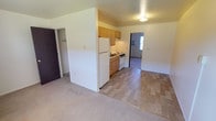 This single-floor unit offers a straightforward, functional layout designed for comfortable everyday living. The open living area features neutral-toned carpet and connects directly to the kitchen, which is partially separated by a dividing wall. The kitchen is equipped with light oak cabinetry providing generous upper and lower storage, a double stainless steel sink set into a light countertop, a white electric coil-burner range, and a white refrigerator. An overhead exhaust fan is positioned above the range for ventilation.

The bedroom features the same neutral carpeting found throughout the living spaces and includes a window with blinds that allows natural light to filter in. A wall-mounted window air conditioning unit provides cooling, while baseboard heating along the exterior wall ensures warmth. A spacious closet with sliding dark-panel doors and built-in rods offers practical storage.

The bathroom is cleanly appointed with a tub and shower combination enclosed by a sliding glass door, a toilet, and a mirrored medicine cabinet with integrated lighting above. Throughout the unit, the clean white walls and simple finishes create a bright, neutral atmosphere ready for personalization.