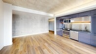 Located in Portland's vibrant inner eastside, this 2-bedroom, 2-bathroom residence offers a compelling blend of industrial-modern design and refined finishes. The open-concept living, dining, and kitchen area is defined by striking exposed concrete ceilings and walls, contrasted by warm hardwood flooring that runs throughout. Floor-to-ceiling windows flood the space with natural light and frame sweeping views of the Portland skyline and Willamette River, with direct access to a private balcony. The kitchen features sleek purple-toned cabinetry, stainless steel appliances, and a geometric tile backsplash, creating a bold yet cohesive aesthetic. Subtle cove lighting along the ceiling adds warmth and dimension to the open living space. Both bedrooms continue the concrete ceiling detail and are carpeted for comfort, with large windows that invite abundant daylight and maintain the expansive city views. Each bedroom is supported by its own dedicated closet. The two bathrooms are clean and functional, finished with modern fixtures, tub-shower combinations, and warm-toned flooring. A hallway connects the private quarters to the main living areas, and an in-unit laundry area adds everyday convenience. The overall ambiance is contemporary and airy, with an industrial edge that reflects Portland's architectural character.