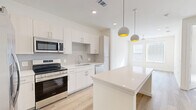 This thoughtfully designed single-floor residence offers a clean, contemporary aesthetic throughout. The open-concept kitchen and living area serves as the heart of the home, anchored by a large white quartz island that doubles as a dining and prep surface. The kitchen is outfitted with white flat-panel cabinetry, a subway tile backsplash, stainless steel appliances including a range, microwave, dishwasher, and French door refrigerator, and a stainless undermount sink. Distinctive globe pendant lights with gold interiors add a warm, modern touch overhead, while light wood-look flooring runs seamlessly throughout the entire space. The bedroom is bright and airy, featuring dual windows with slatted blinds that invite generous natural light and a ceiling fan for added comfort. Two closets provide ample storage. The bathroom is elegantly appointed with a long floating white vanity, a wide framed mirror, a tall storage tower, and a glass-enclosed shower with tile surround. A dedicated laundry area adds everyday convenience. The overall palette of crisp whites, soft neutrals, and warm wood tones creates a cohesive, modern ambiance that feels both stylish and functional.