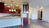 This single-floor residence offers a thoughtfully designed open-concept layout that seamlessly connects the kitchen and living areas, creating a spacious and functional environment. The kitchen is a standout feature, showcasing rich dark cherry wood cabinetry that extends from counter height to the ceiling, maximizing storage while adding warmth and elegance to the space. Stainless steel appliances, including a gas range, microwave, and refrigerator, provide a modern touch that complements the cabinetry's deep tones. A peninsula counter with a built-in sink serves as both a practical workspace and a natural divider between the kitchen and the adjoining living area, ideal for casual dining or entertaining. Pendant lighting hangs above the peninsula, adding focused illumination and a decorative element to the kitchen zone. A distinctive built-in column cabinet with glass-front upper doors and solid lower doors adds architectural interest and additional storage between spaces. The flooring throughout is a rich, dark hardwood that anchors the interior with a sense of continuity and sophistication. Recessed lighting across the ceiling ensures the space is well-lit, while the open corridor leading to additional rooms suggests a well-planned floor plan designed for comfortable everyday living.