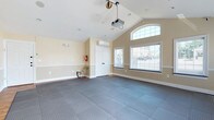 This single-floor multipurpose space is designed for flexibility and function, offering an open, airy layout well-suited for a variety of activities and gatherings. The standout architectural feature is the soaring vaulted ceiling, which creates a sense of generous volume and height throughout the room. A ceiling-mounted projector pairs with a large wall-mounted projection screen, making the space ideal for presentations, screenings, or group events.

Natural light pours in through multiple large windows fitted with horizontal blinds, including a striking arched transom window that adds an elegant architectural touch. A set of French doors provides an additional point of entry and further brightens the interior with views to the outdoors. The flooring consists of interlocking foam or rubber mat tiles covering much of the open floor area, with hardwood flooring visible along the perimeter — a practical combination suited for fitness, yoga, martial arts, or similar activities.

The walls feature a warm neutral tone with a chair-rail detail, contributing to a clean and welcoming ambiance. A wall-mounted mini-split HVAC unit ensures climate control year-round. Recessed ceiling speakers and lighting enhance the space's functionality. Safety features include a fire extinguisher and clearly marked emergency exits, reflecting a thoughtful and code-compliant design.