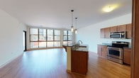 This single-floor residence features an open-concept layout that seamlessly connects the kitchen, dining, and living areas. The living space is anchored by warm hardwood flooring that runs throughout the main area, complemented by crisp white walls and natural light pouring in through a large bank of floor-to-ceiling windows that frame urban views of the surrounding cityscape.

The kitchen is well-appointed with rich wood cabinetry extending to the ceiling, dark granite countertops, and a full suite of stainless steel appliances including a refrigerator, electric range, and built-in microwave. A central island with a granite surface and undermount sink doubles as a breakfast bar, with stylish pendant lighting suspended above.

The bedrooms are carpeted for comfort, each offering generous closet space. One bedroom features an expansive window wall with views of an outdoor terrace area, filling the room with abundant natural light.

The bathroom includes a granite-topped vanity with wood cabinetry, a full soaking tub with shower, white tile flooring, and a window for natural ventilation. Additional storage is provided by a walk-in closet near the entryway. The overall ambiance is clean, modern, and functional.