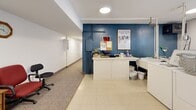 Located at 405 Tenth Ave N in South Saint Paul, Minnesota, this single-level utility space offers a practical and well-organized layout designed for functional everyday use. The space is divided into distinct zones, each serving a specific purpose while maintaining a cohesive, no-frills aesthetic.

The laundry area forms the core of the space, equipped with washing machines and dryers arranged along white cabinetry. Exposed overhead piping and ventilation ducts reflect the utility-forward design. A window allows natural light to filter in, providing views of surrounding greenery and keeping the area from feeling entirely enclosed. The flooring here is a light-toned vinyl tile, easy to maintain and durable for high-use conditions.

Adjacent to the laundry zone is a counter workspace set against a bold blue accent wall, which adds a touch of visual contrast to the otherwise neutral palette. This area includes a built-in counter with cabinet storage below, suitable for sorting, folding, or administrative tasks.

A carpeted seating nook transitions the space into a quieter area, furnished with office-style seating and a small side cabinet. Overhead circular flush-mount lighting illuminates each section consistently throughout the space.

A dedicated storage wall features multiple panel-door compartments, offering organized, lockable storage options. The combination of tile and carpet flooring delineates functional zones clearly, making this a versatile and efficient utility space.