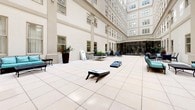 This open-air atrium courtyard is a refined and versatile outdoor space nestled within the embrace of a multi-story building with a classic, cream-colored facade punctuated by rows of tall, evenly spaced windows. The expansive floor is laid with large, light-toned stone tiles that lend a clean and polished aesthetic to the entire space.

Multiple seating arrangements are thoughtfully distributed throughout the courtyard, each designed to accommodate different needs. Deep blue cushioned armchairs and loveseats with wrought-iron frames are grouped around low coffee tables, creating comfortable conversation areas. Teal-cushioned chaise lounges with accompanying side tables offer a more leisurely setting, ideal for relaxing. Compact bistro-style tables and chairs provide additional intimate gathering spots.

Potted tropical plants and tall palms are positioned at various points around the perimeter, adding lush greenery and a vibrant sense of life to the otherwise architectural setting. Large planter boxes with flowering plants contribute seasonal color and visual interest.

The courtyard also accommodates recreational activities, with cornhole boards set up on the open tiled expanse, making the space adaptable for both relaxed socializing and light-hearted events. A glass-enclosed interior space is accessible from one end of the courtyard, extending the usable area. Access doors with exit signage are positioned along the building walls, ensuring easy connectivity throughout.