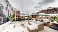 Located atop 267 Main St in downtown New Britain, Connecticut, this rooftop terrace offers an elevated outdoor experience defined by contemporary design and urban flair. The expansive open-air space is finished with large-format light tile flooring throughout, lending a clean, modern aesthetic that unifies the entire terrace. Multiple seating arrangements feature woven rattan furniture with plush white cushions, configured to accommodate both intimate gatherings and larger social events. Large cantilever umbrellas are positioned strategically across the terrace, providing shade while maintaining an open, airy atmosphere. A defining feature of the space is a bold, large-scale mural depicting stylized portrait figures rendered in black, white, and pink tones — a dramatic artistic focal point that anchors one end of the terrace. Raised planter boxes line portions of the perimeter railing, filled with colorful seasonal flowers and tropical foliage that add vibrancy and a sense of greenery to the urban setting. The terrace offers unobstructed panoramic views of the New Britain skyline, with historic brick buildings and rolling hills visible in the distance. High-top table seating provides additional options for guests. The overall ambiance blends sophisticated outdoor living with an artistic, energetic character unique to this rooftop venue.